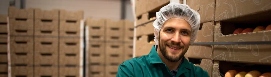 Un jeune homme en charlotte devant des cageots de pommes dans une usine.