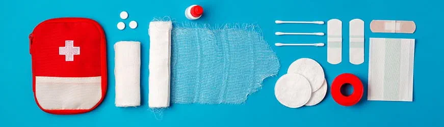 Trousse de premiers secours rouge avec une croix blanche. À côté, des bandes, des cotons tiges, des pansements, du sparadrap, du désinfectant et des pilules.