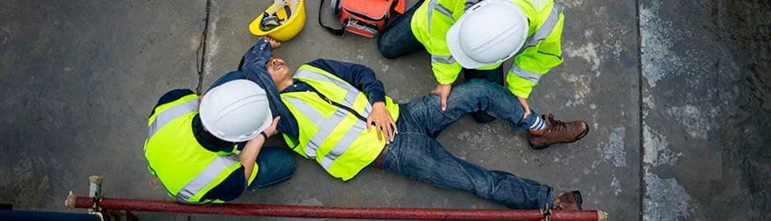 Scène d’accident sur un chantier. Deux personnes portent secours à un homme allongé à terre. Les trois individus portent un gilet jaune à bandes réfléchissantes Scène d’accident sur un chantier. Deux personnes portent secours à un homme allongé à terre. Les trois individus portent un gilet jaune à bandes réfléchissantes