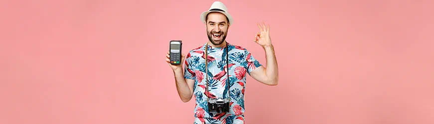 Jeune homme de face sur fond rose. Il porte un chapeau de paille, une chemise à motifs exotiques et en appareil photo en bandoulière. Il brandit d’une main un terminal de paiement en souriant et, de l’autre, il fait le signe OK Jeune homme de face sur fond rose. Il porte un chapeau de paille, une chemise à motifs exotiques et en appareil photo en bandoulière. Il brandit d’une main un terminal de paiement en souriant et, de l’autre, il fait le signe OK