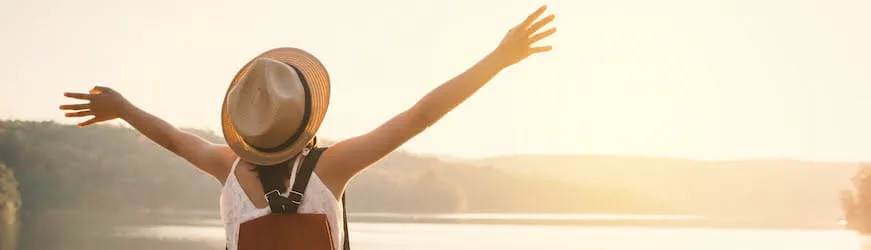 Une salariée, face à la mer et les bras levés, portant un chapeau de paille et un sac à dos, profite de son congé sabbatique