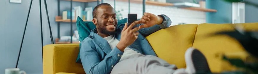 Jeune homme allongé dans son canapé, souriant, regardant son téléphone portable. 
