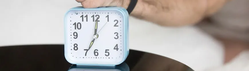 Un jeune homme au lit qui éteint son réveil indiquant 7h : l’heure de se lever pour ne pas arriver en retard au travail !