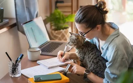 Une salarié·e en télétravail, qui écrit assise à son bureau avec son chat sur les genoux. Une salarié·e en télétravail, qui écrit assise à son bureau avec son chat sur les genoux.