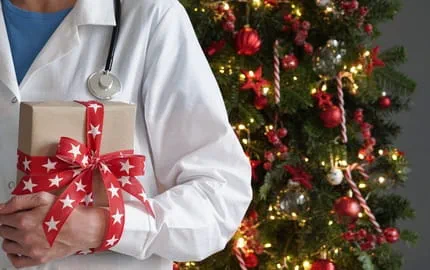Un soignant tenant un cadeau devant un sapin de Noël illuminé, illustrant un Secret-Santa dans le secteur de la santé. Un soignant tenant un cadeau devant un sapin de Noël illuminé, illustrant un Secret-Santa dans le secteur de la santé.