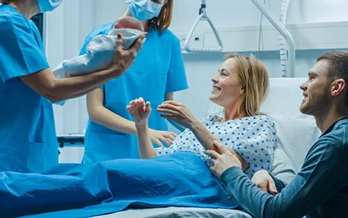 Une soignante présente un nouveau-né à une mère souriante et à son partenaire dans une salle d'accouchement d'hôpital Une soignante présente un nouveau-né à une mère souriante et à son partenaire dans une salle d'accouchement d'hôpital