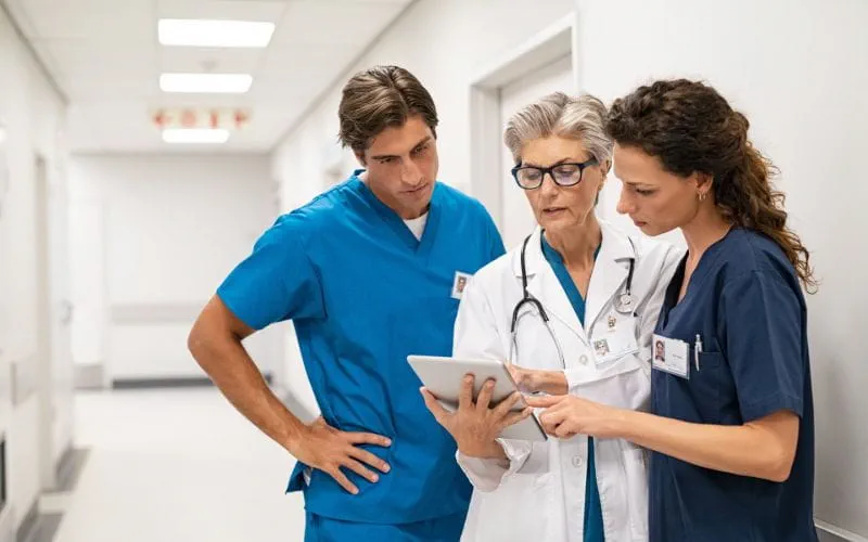 Trois professionnels de la santé, deux femmes et un homme, se tiennent dans un couloir d'hôpital et examinent ensemble une tablette Trois professionnels de la santé, deux femmes et un homme, se tiennent dans un couloir d'hôpital et examinent ensemble une tablette