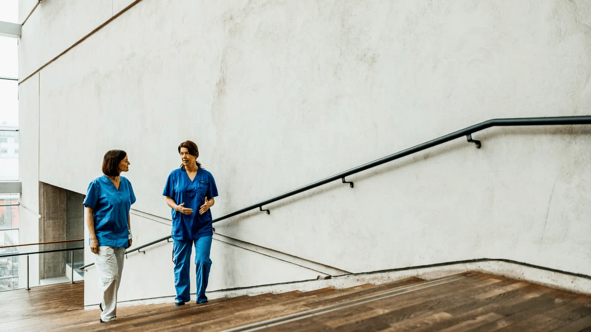 Two healthcare professionals in blue scrubs walk together and converse on a wooden staircase in a bright, modern hospital corridor. Two healthcare professionals in blue scrubs walk together and converse on a wooden staircase in a bright, modern hospital corridor.