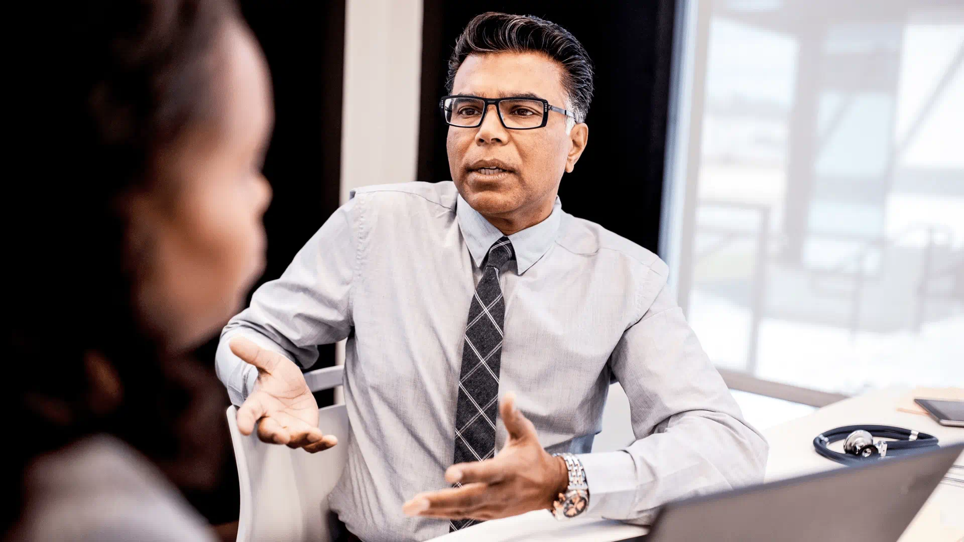 A man wearing glasses, a dress shirt, and a tie gestures while speaking with an individual seated across from him. A man wearing glasses, a dress shirt, and a tie gestures while speaking with an individual seated across from him.