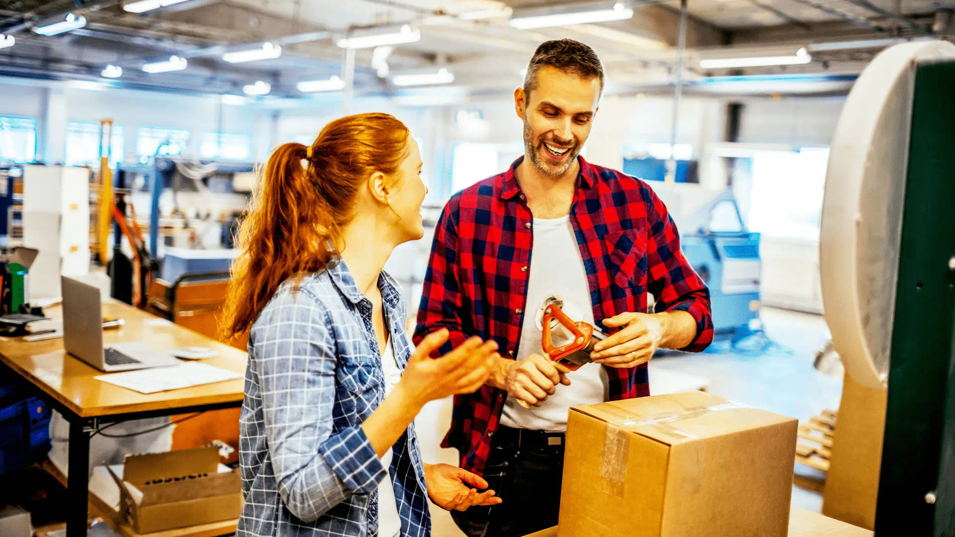 Two coworkers smiling and talking in a workshop, with the man holding a handheld power tool beside a large cardboard box. Two coworkers smiling and talking in a workshop, with the man holding a handheld power tool beside a large cardboard box.