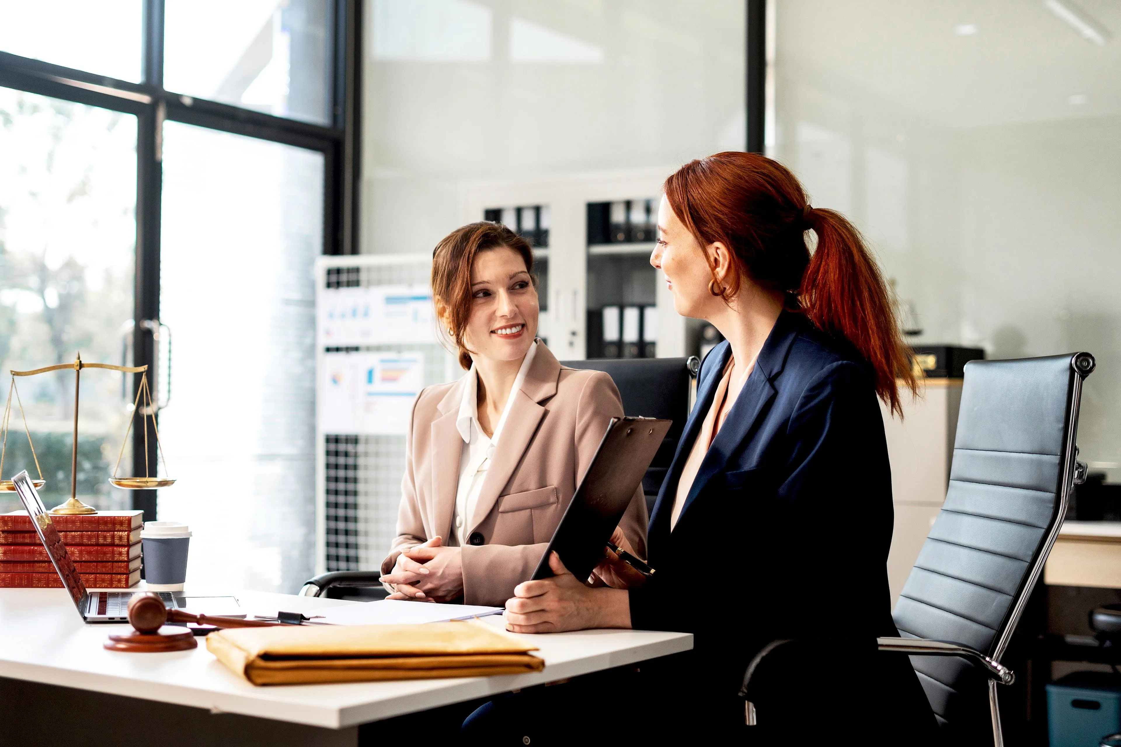 two women dressed in suits sitting at a desk in a work office, smiling two women dressed in suits sitting at a desk in a work office, smiling
