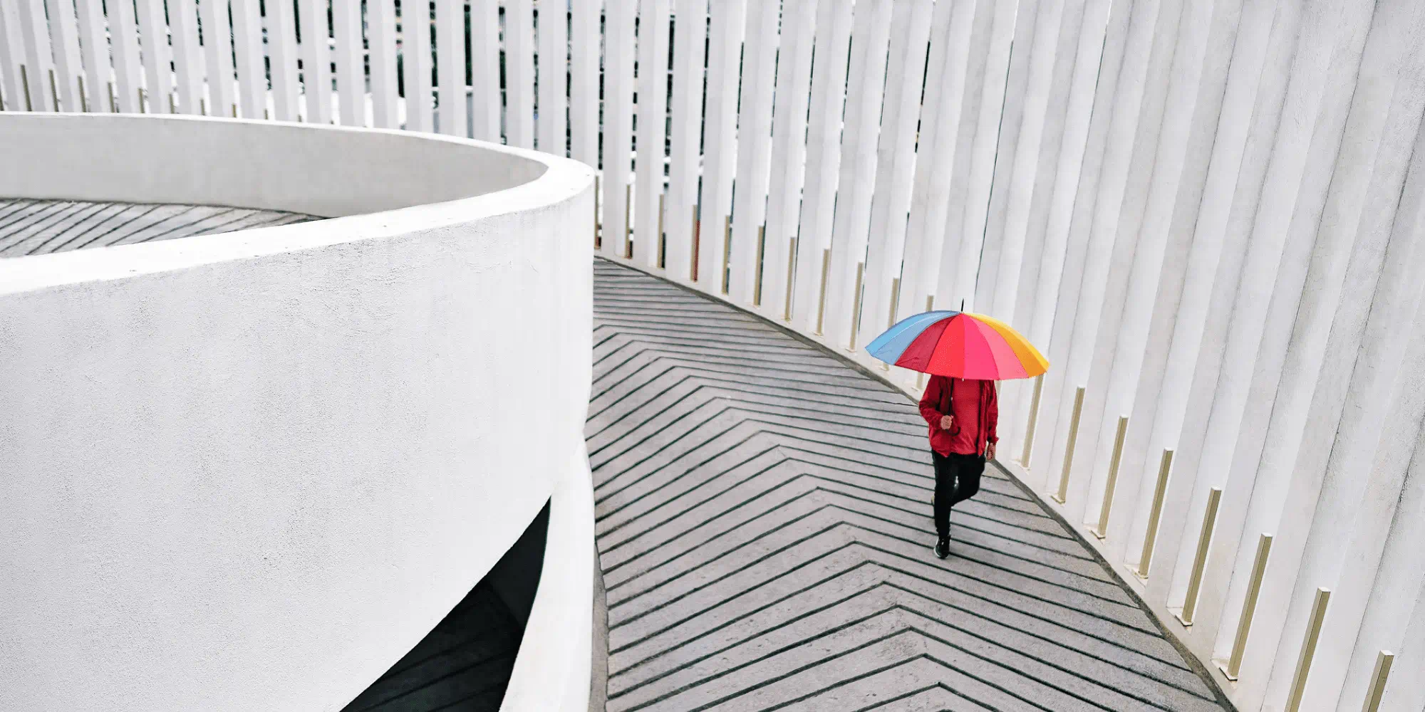 A person carrying a colourful umbrella walks up a car park ramp between bright, modern walls. A person carrying a colourful umbrella walks up a car park ramp between bright, modern walls.