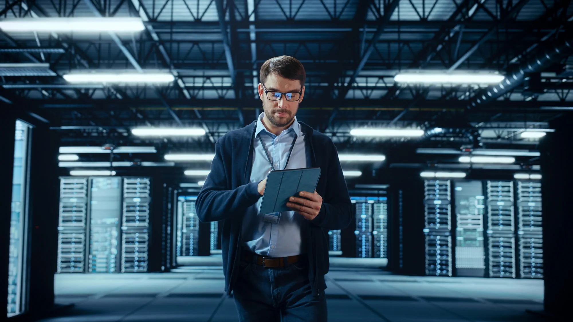 Male IT specialist walks between rows of operational server racks in a data center. Engineer uses a tablet computer for maintenance. Concept for cloud computing, artificial intelligence, and cybersecurity. Male IT specialist walks between rows of operational server racks in a data center. Engineer uses a tablet computer for maintenance. Concept for cloud computing, artificial intelligence, and cybersecurity.