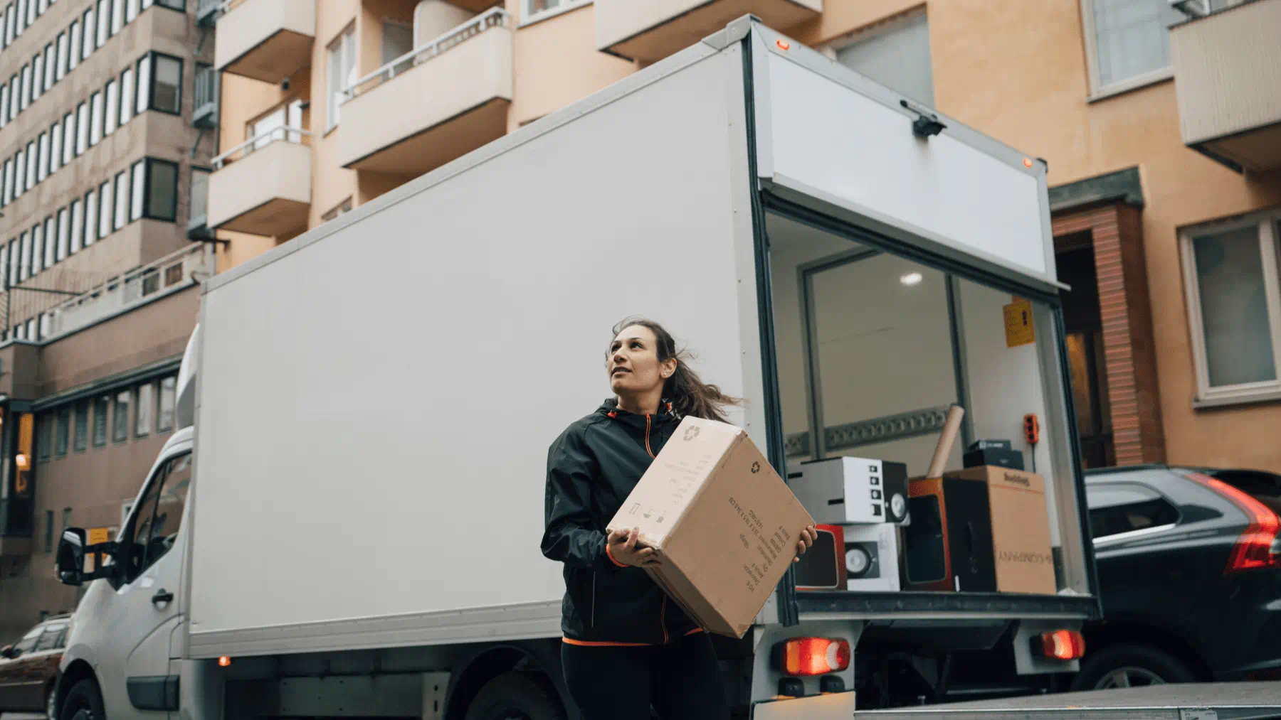 Person carrying a cardboard box from the back of a white delivery truck parked on a city street. Apartment buildings and parked cars are visible in the background, and additional moving boxes and household items are loaded inside the truck. Person carrying a cardboard box from the back of a white delivery truck parked on a city street. Apartment buildings and parked cars are visible in the background, and additional moving boxes and household items are loaded inside the truck.