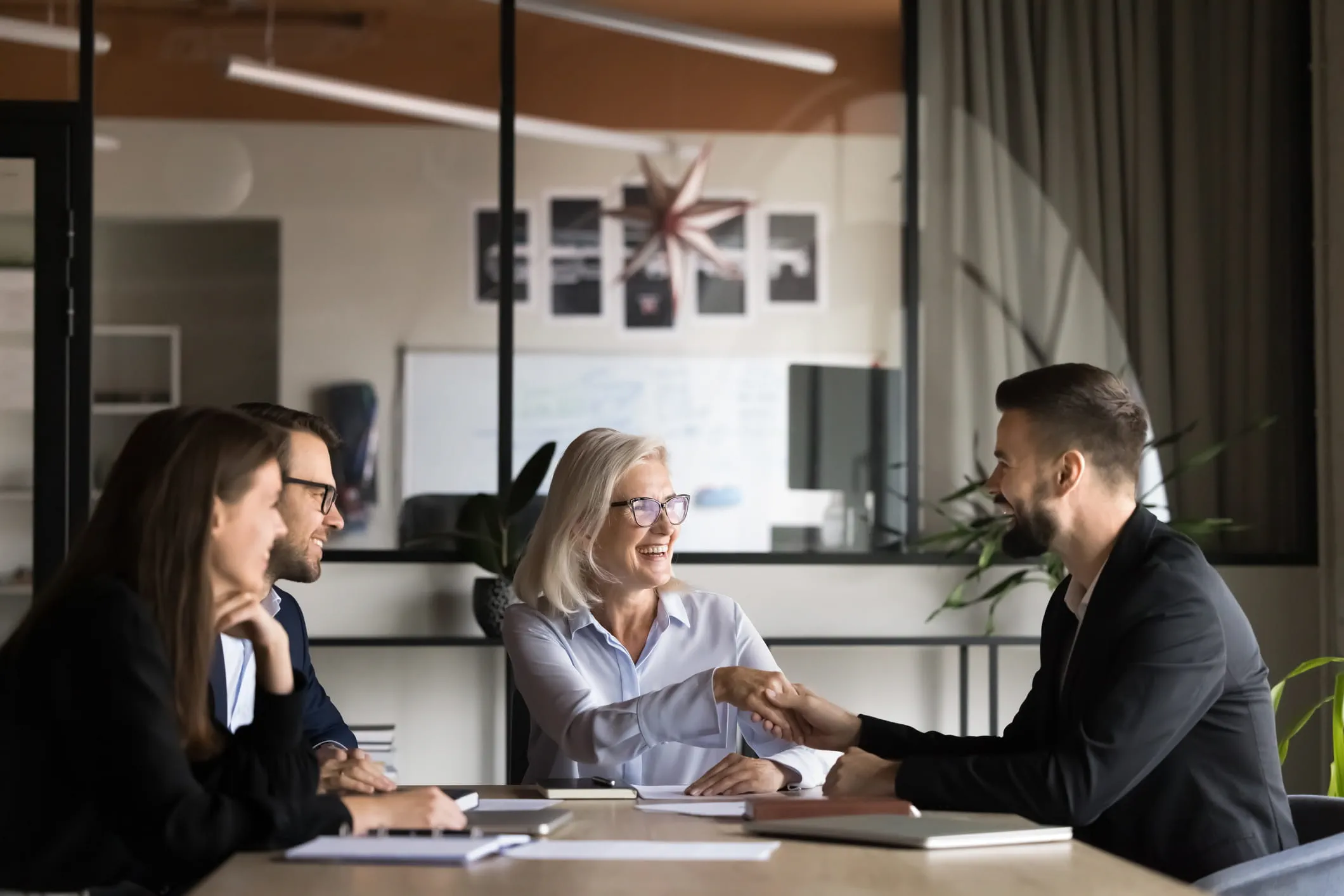 Four people sit at a conference table in a modern office; two people shake hands across the table, with documents and notebooks placed on the table.