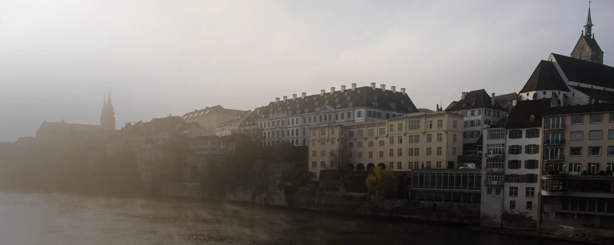 Foggy riverside scene of historic buildings along the water in a European city, with church towers partially obscured by mist.