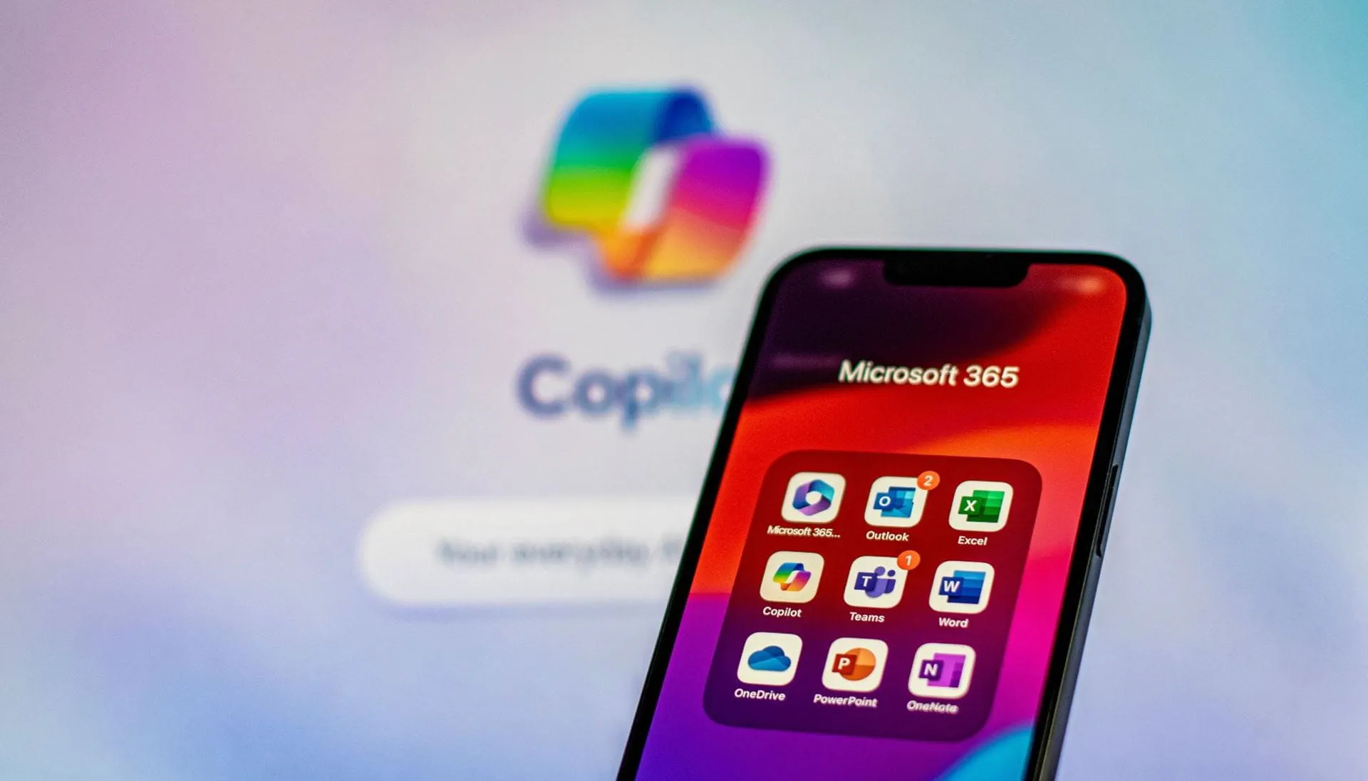 Sullo schermo di uno smartphone viene visualizzata una cartella intitolata "Microsoft 365". All'interno della cartella ci sono diverse icone di app: Amministratore di Microsoft 365 Outlook (con due badge di notifica) Eccellere Copilota Teams (con un badge di notifica) Parola OneDrive Punto di forza OneNote Sullo sfondo sfocato, c'è un grande logo Microsoft Copilot con la parola "Copilot" parzialmente visibile sotto di esso. Lo sfondo presenta una morbida sfumatura di colori pastello, che passano dal viola al blu.