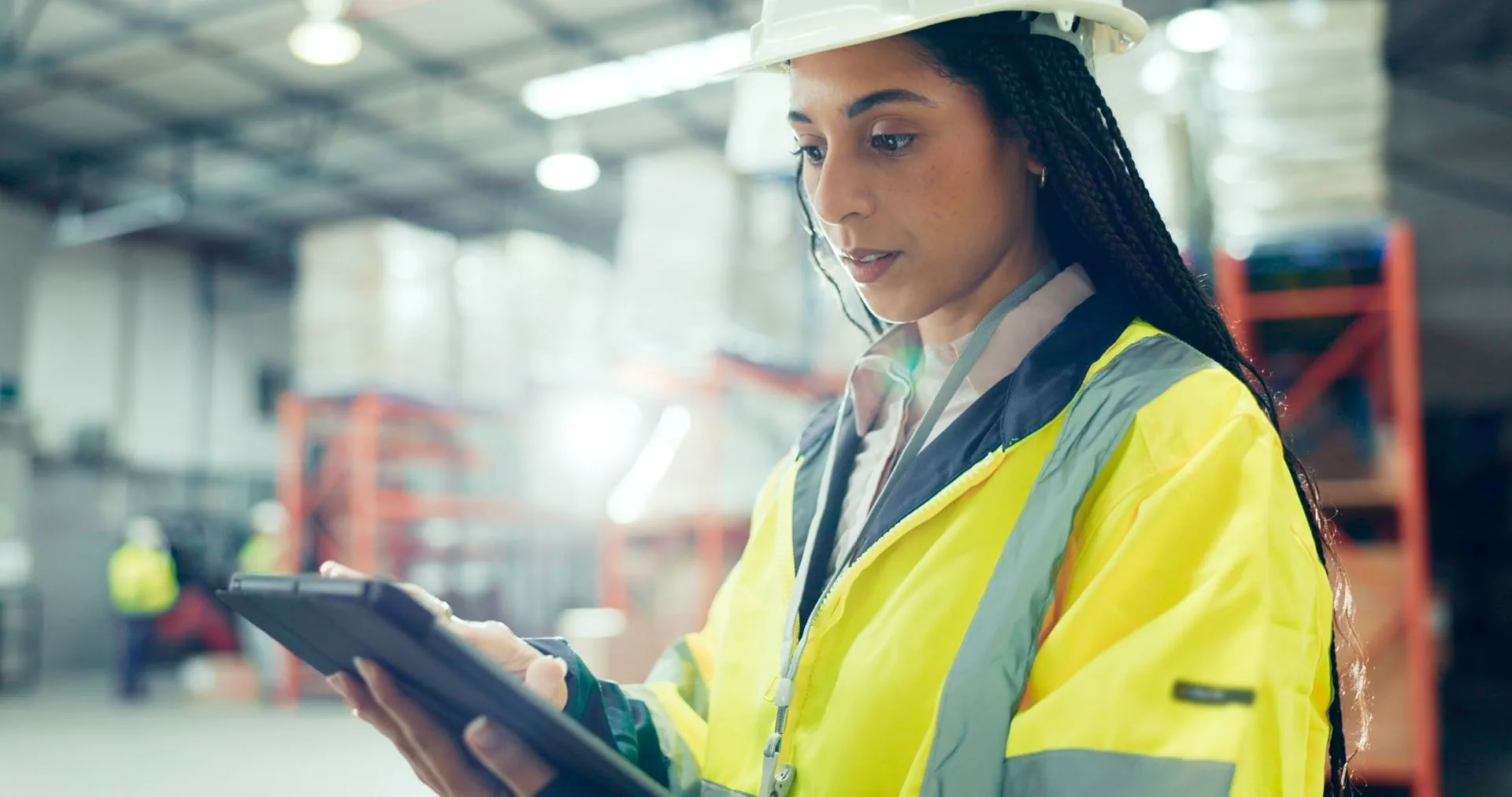 Una donna con una giacca di sicurezza ad alta visibilità e un casco bianco si trova in un magazzino mentre guarda un tablet. Sullo sfondo, leggermente sfocato, si vedono scaffali e lavoratori, suggerendo un ambiente industriale movimentato.