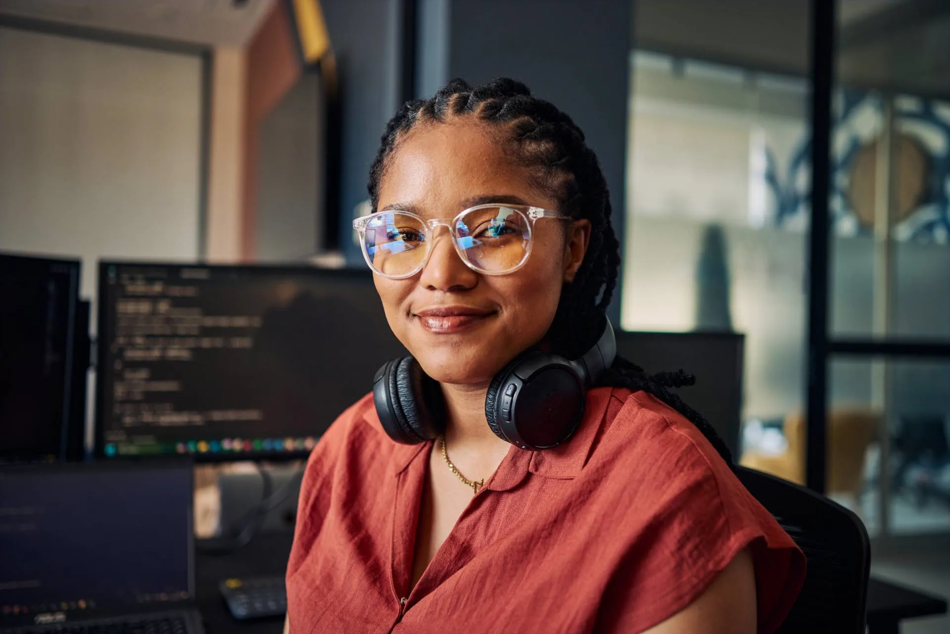 Foto einer lächelnden Frau mit geflochtenen Haaren und Brille an einem Arbeitsplatz. Sie trägt ein rotes Oberteil und Kopfhörer um den Hals; im Hintergrund sind Monitore mit unscharfem Code in einem modernen Büro zu sehen. Foto einer lächelnden Frau mit geflochtenen Haaren und Brille an einem Arbeitsplatz. Sie trägt ein rotes Oberteil und Kopfhörer um den Hals; im Hintergrund sind Monitore mit unscharfem Code in einem modernen Büro zu sehen.