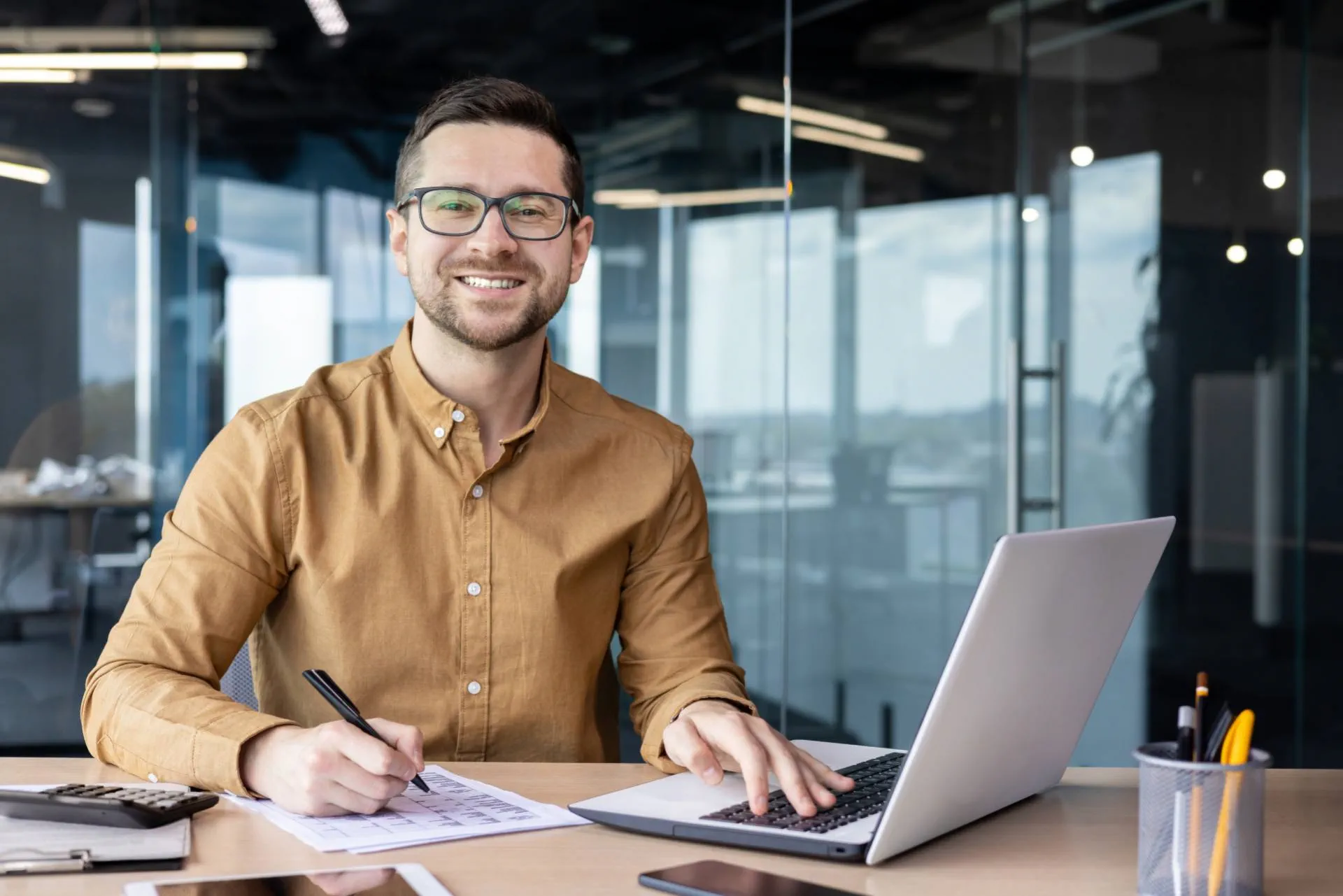 Mann sitzt in einem modernen Büro am Schreibtisch und arbeitet am Laptop. Er lächelt in die Kamera. Auf dem Tisch liegen Rechner, Dokumente und Stifte. Mann sitzt in einem modernen Büro am Schreibtisch und arbeitet am Laptop. Er lächelt in die Kamera. Auf dem Tisch liegen Rechner, Dokumente und Stifte.