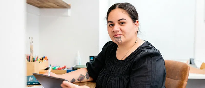 Person with Māori tāmoko tattoos working at a desk with a pen and tablet in an office setting Person with Māori tāmoko tattoos working at a desk with a pen and tablet in an office setting