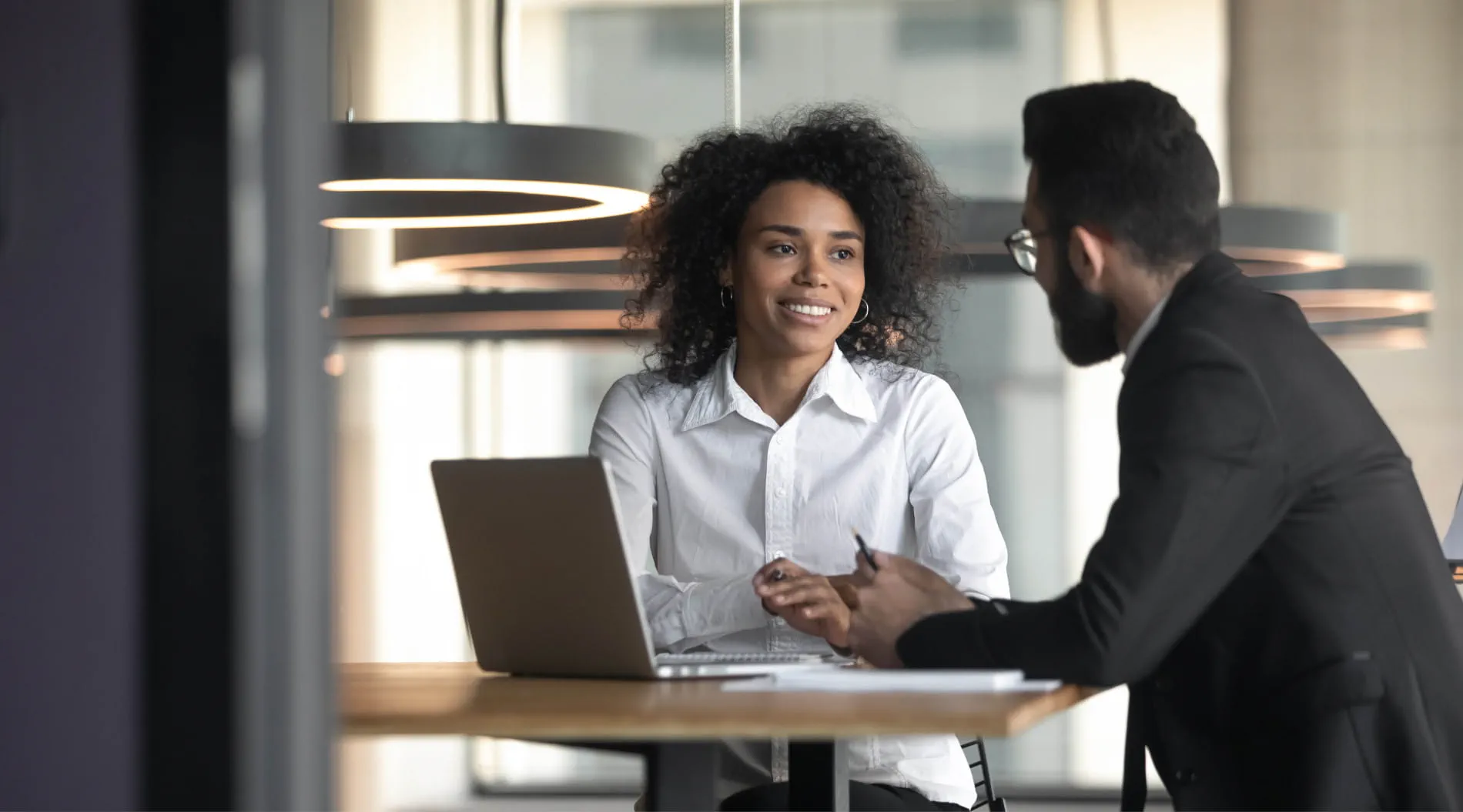 Two people in professional attire having a discussion at a table with a laptop in an office setting Two people in professional attire having a discussion at a table with a laptop in an office setting