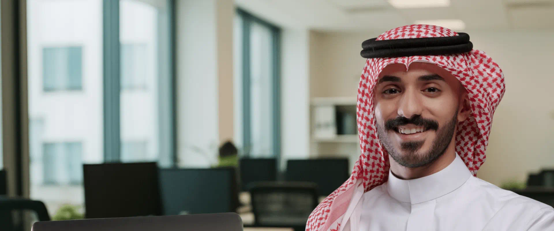 Smiling Saudi man in traditional attire working on a laptop in a modern office environment Smiling Saudi man in traditional attire working on a laptop in a modern office environment