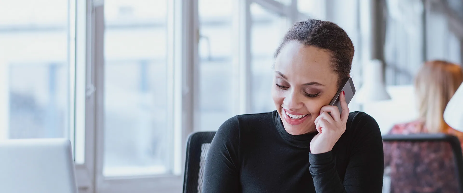 A smiling professional woman in a black top engages in a phone call in a bright, modern office with large windows. A smiling professional woman in a black top engages in a phone call in a bright, modern office with large windows.