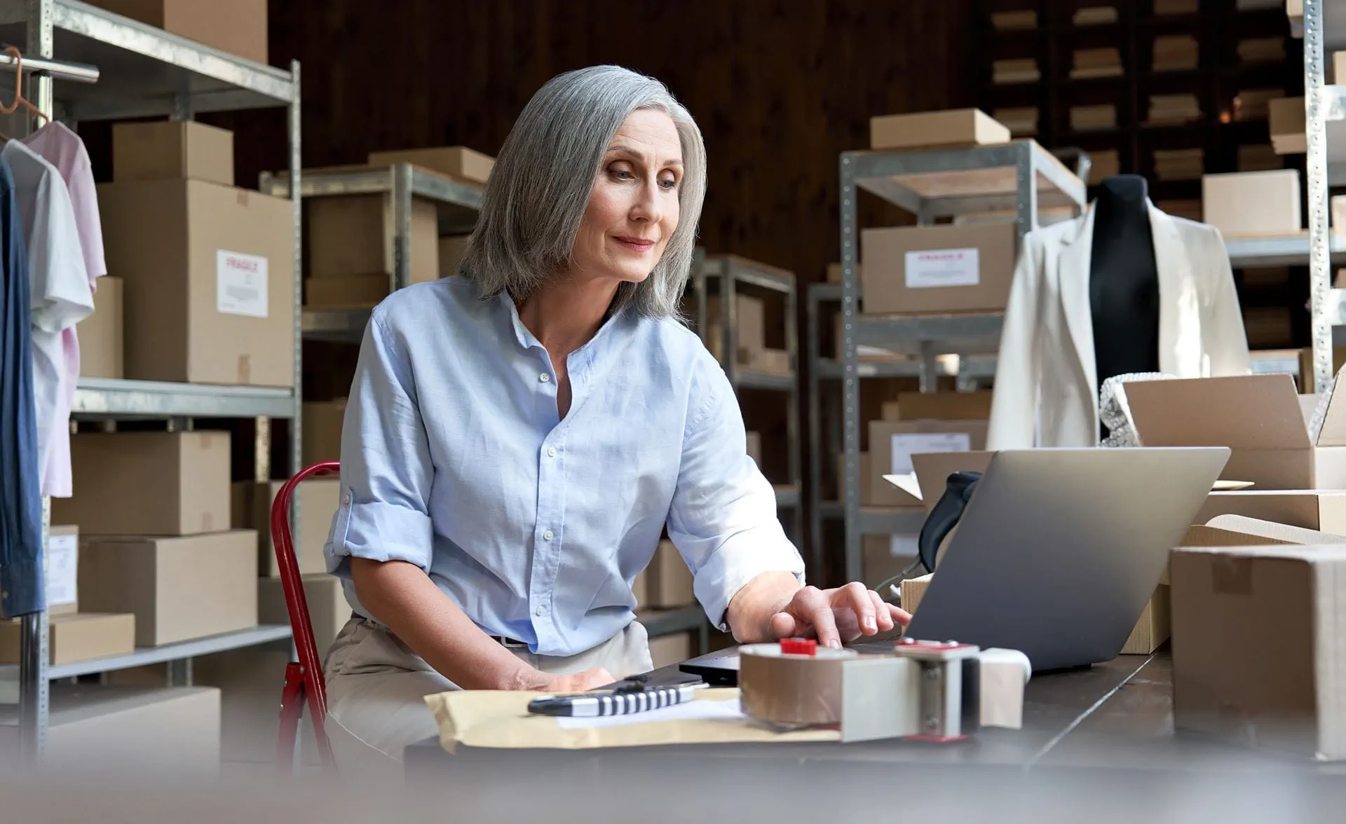 A woman in her late 40s works at a computer in a warehouse office, managing administrative tasks and paperwork in a logistics environment. A woman in her late 40s works at a computer in a warehouse office, managing administrative tasks and paperwork in a logistics environment.