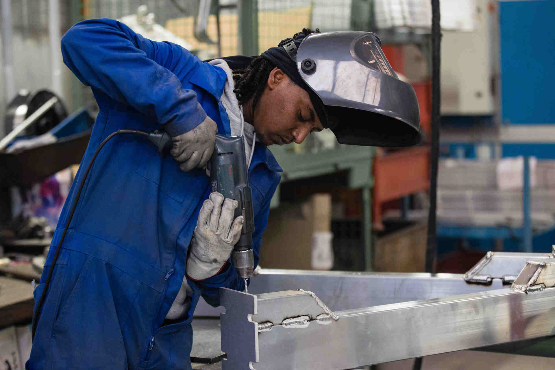 Worker in a blue coverall and protective helmet using a power tool to work on a metal piece in an industrial setting with machinery in the background. Worker in a blue coverall and protective helmet using a power tool to work on a metal piece in an industrial setting with machinery in the background.