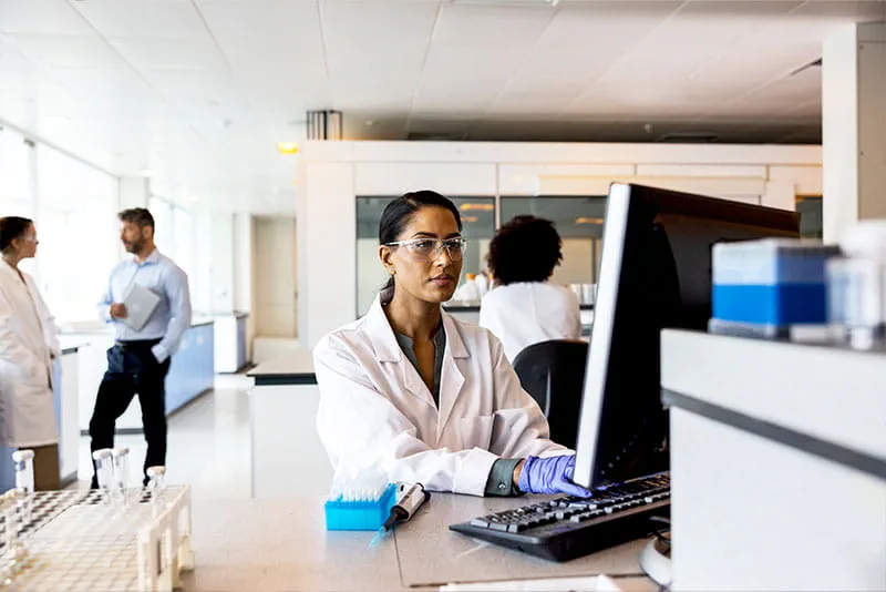 Scientist working at a computer in a modern laboratory while colleagues collaborate in the background.