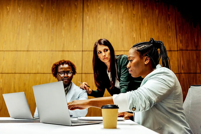 Three colleagues collaborating in a meeting room, reviewing work on a laptop while discussing ideas around a table. Three colleagues collaborating in a meeting room, reviewing work on a laptop while discussing ideas around a table.