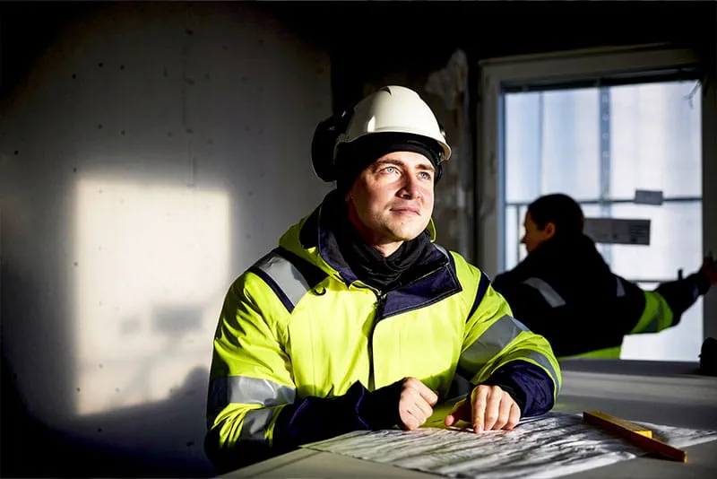 Construction worker wearing a high-visibility jacket and hard hat reviewing building plans indoors with a colleague in the background. Construction worker wearing a high-visibility jacket and hard hat reviewing building plans indoors with a colleague in the background.