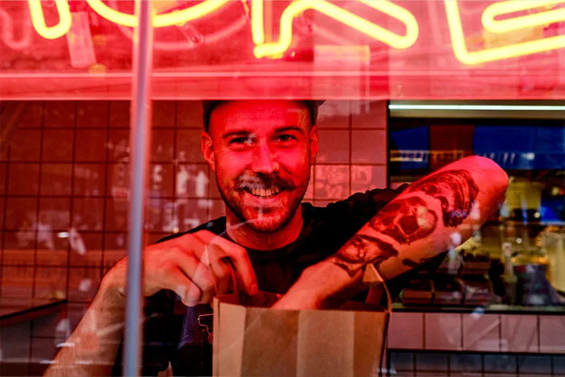A smiling worker behind a counter packing an order, illuminated by red neon lighting.
