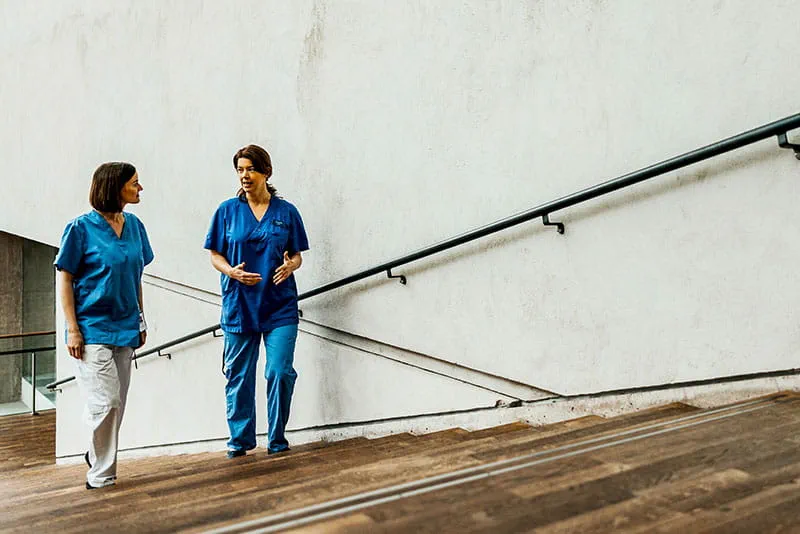 Two healthcare professionals in scrubs walking up stairs, talking and collaborating in a clinical setting. Two healthcare professionals in scrubs walking up stairs, talking and collaborating in a clinical setting.