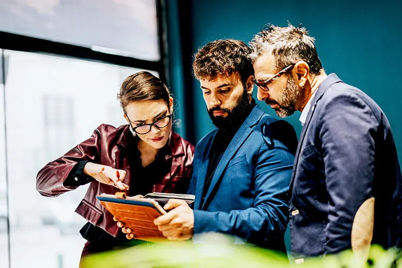 Three professionals reviewing information together on a tablet during a discussion in a modern office. Three professionals reviewing information together on a tablet during a discussion in a modern office.