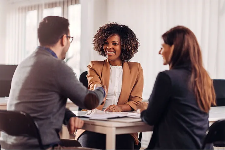 Three business professionals sitting at a table in an office, with one person smiling and shaking hands with another, indicating a successful meeting or agreement. Three business professionals sitting at a table in an office, with one person smiling and shaking hands with another, indicating a successful meeting or agreement.