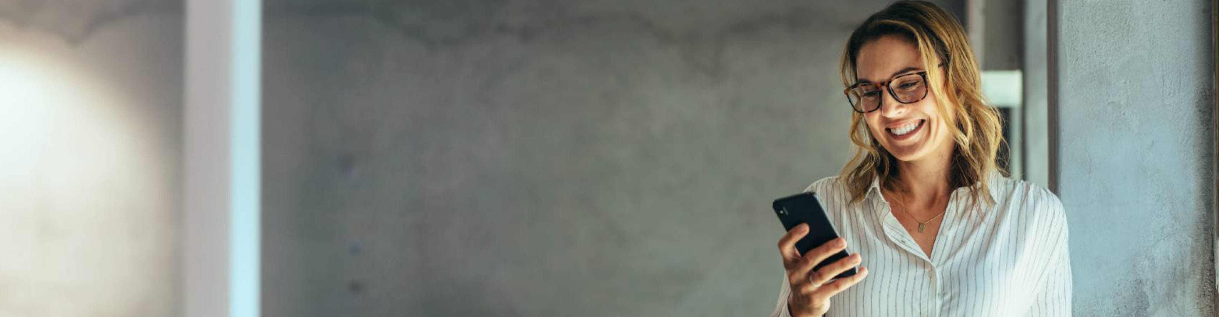 Une femme blonde en chemise blanche sourit devant son téléphone.