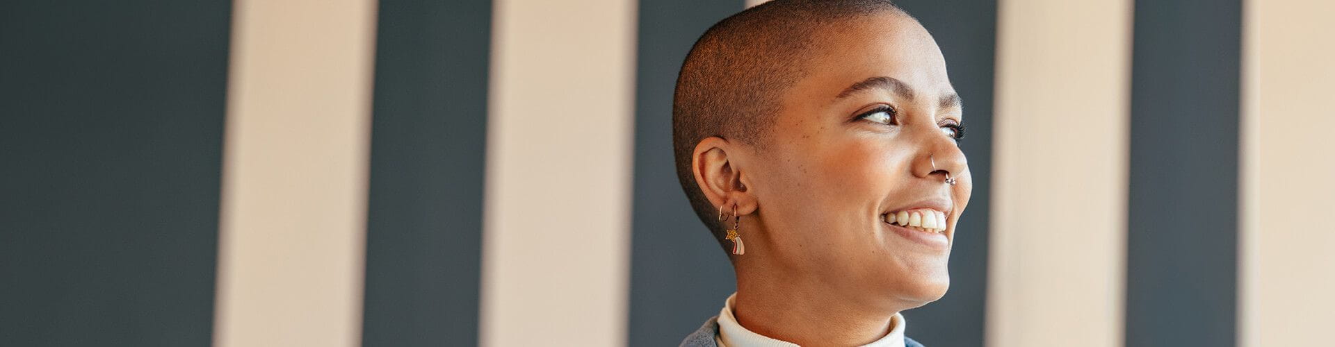 Portrait en profil d'une jeune femme souriante avec des cheveux courts et des piercings devant un mur à rayures