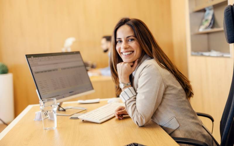 Une jeune femme brune aux cheveux longs souriante travaille sur son ordinateur dans un bureau
