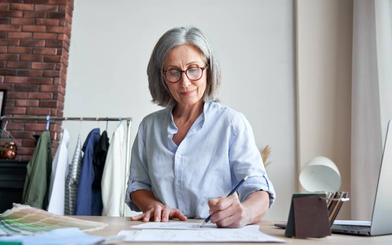 Une femme aux cheveux gris et lunettes dessinant sur du papier à un bureau avec des vêtements suspendus en arrière-plan
