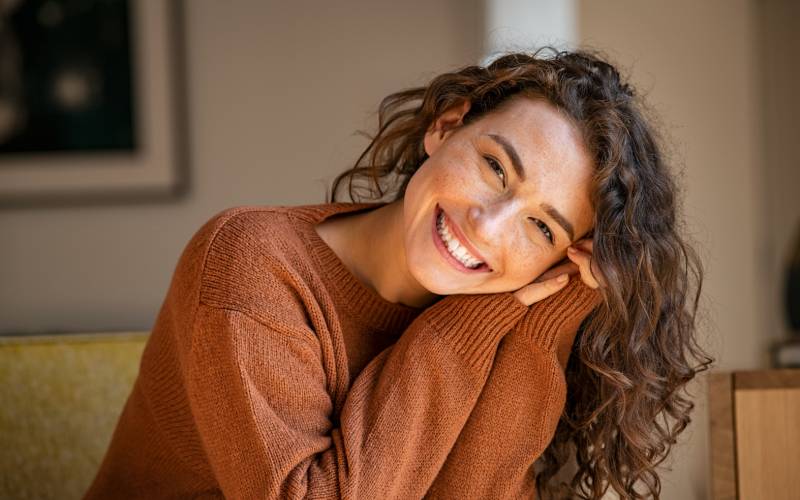 Une femme joyeuse avec un pull marron joint ses deux mains et rigole en regardant la caméra