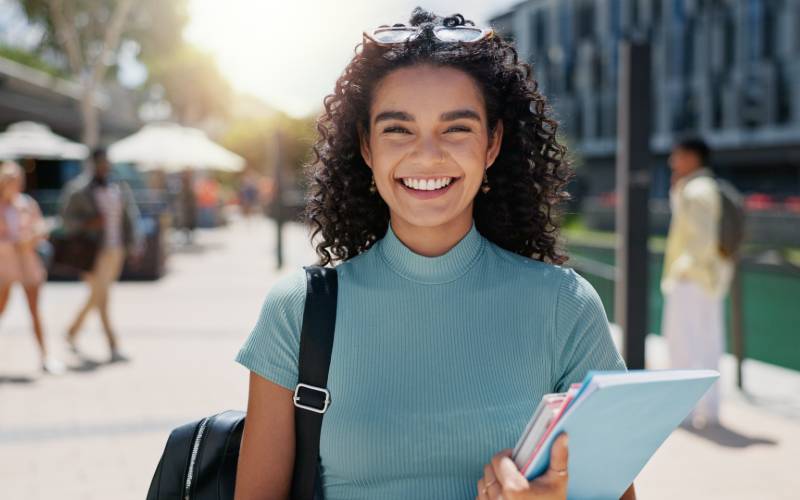 Une étudiante souriante avec des lunettes de soleil sur la tête, portant un sac à dos et des livres sur un campus universitaire ensoleillé