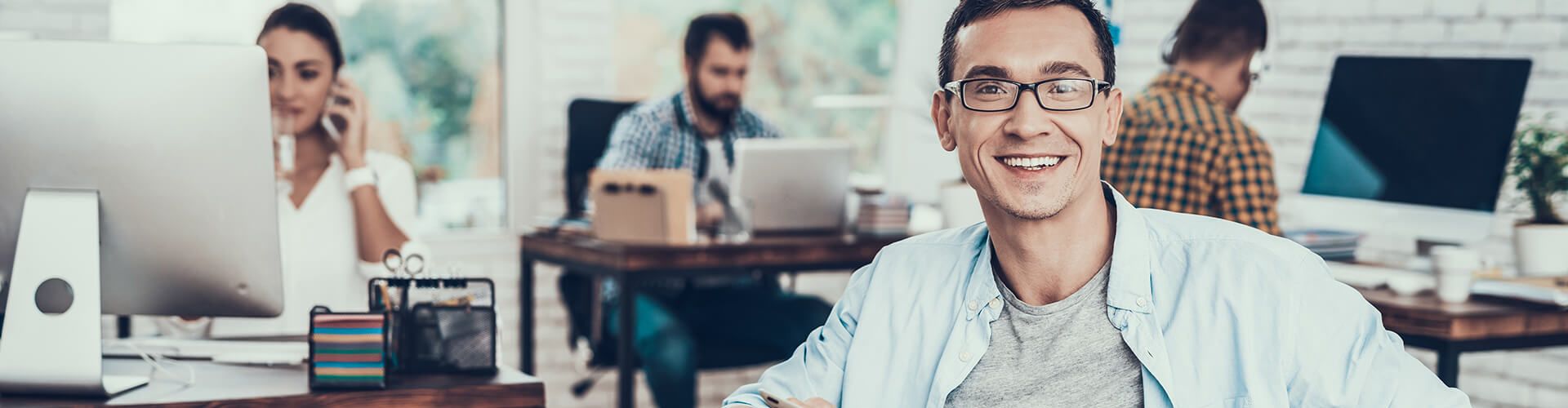 Un jeune homme souriant avec des lunettes dans un bureau de style décontracté avec d'autres personnes travaillant en arrière-plan