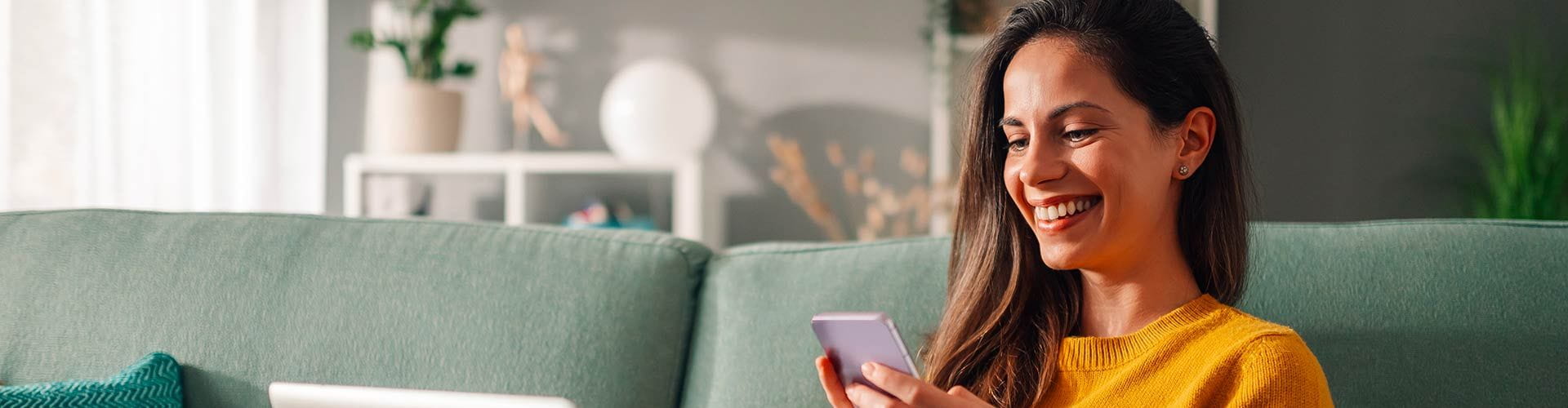 Une jeune femme souriante assise sur un canapé vert clair, regardant son téléphone portable