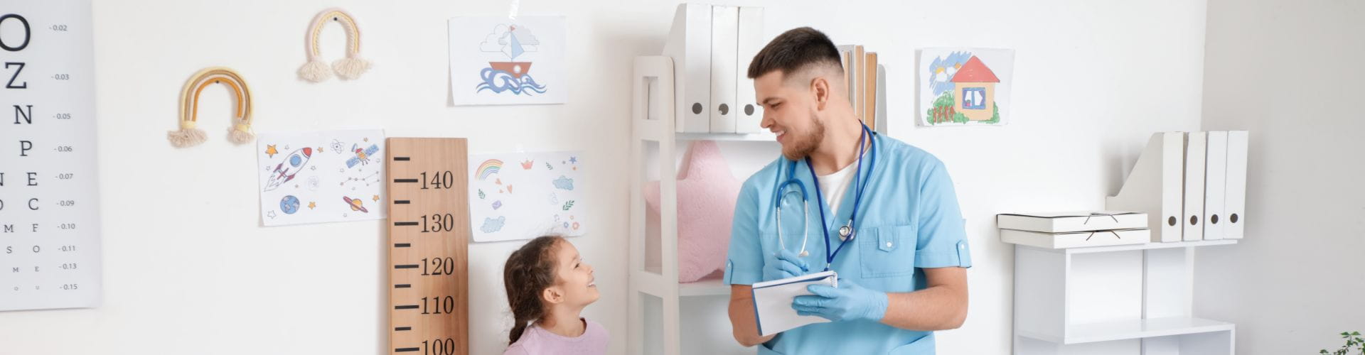 Un pédiatre mesure la taille d'une jeune fille avec des dessins d’enfants accrochés sur le mur