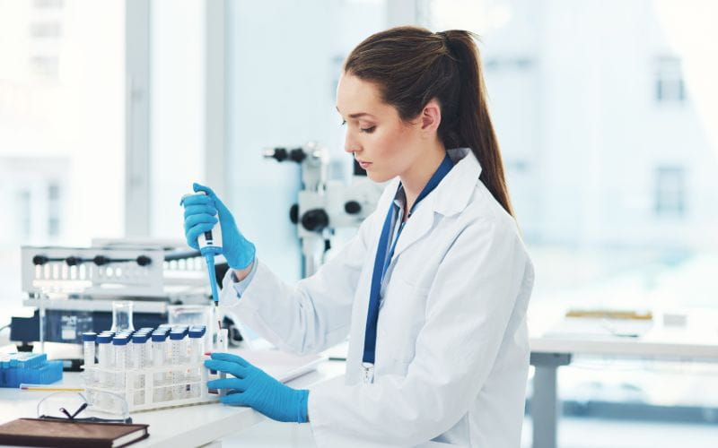 Technicienne de laboratoire en blouse blanche manipulant une pipette pour effectuer des analyses dans un environnement scientifique, entourée de tubes à essai.