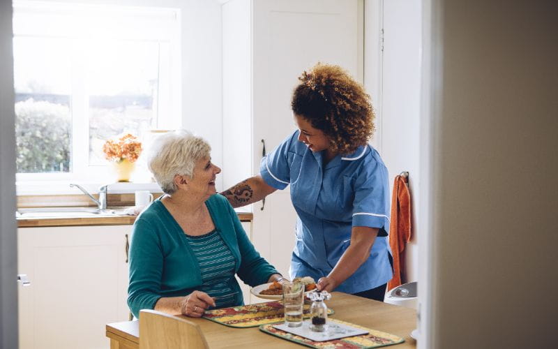 Aide à domicile souriante servant un repas à une personne âgée assise à table dans une cuisine lumineuse, illustrant un moment de bienveillance et d'accompagnement quotidien.