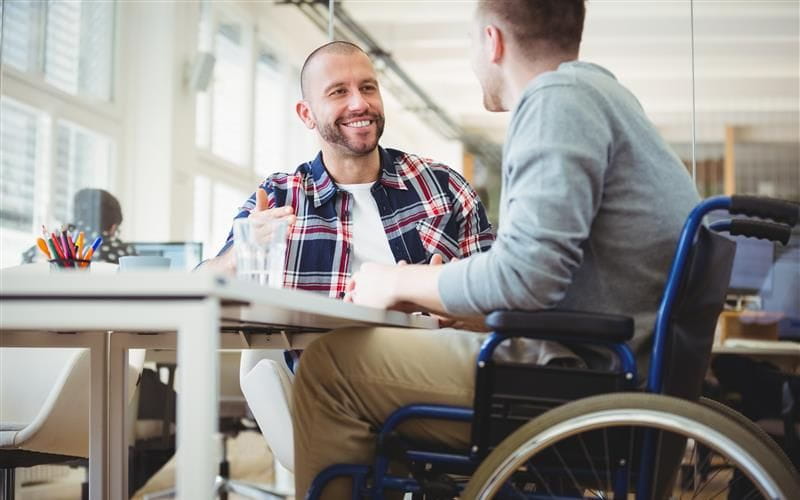 Un homme souriant et barbu, vêtu d'une chemise à carreaux, discute avec une personne en fauteuil roulant