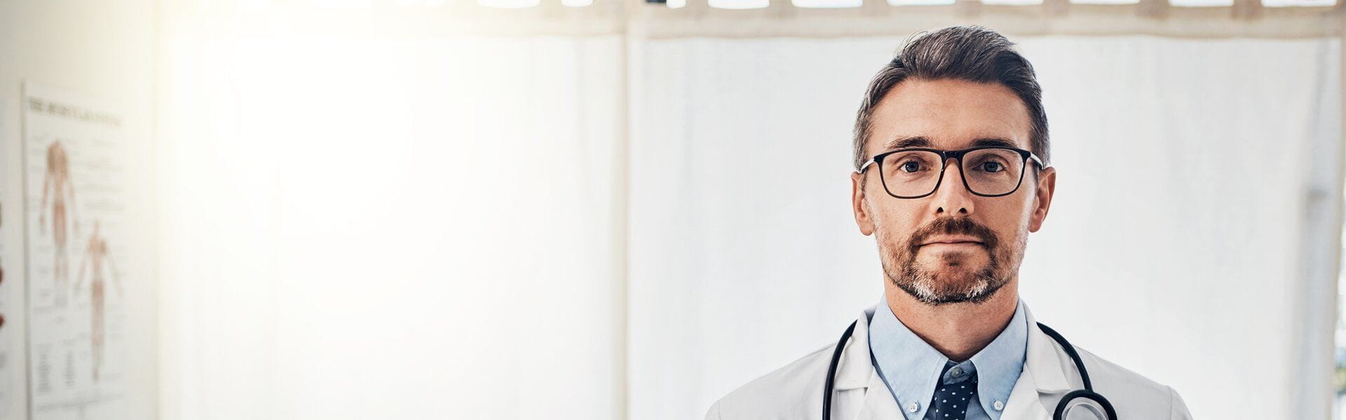 Un médecin avec des lunettes portant un stéthoscope et une blouse, regarde la caméra dans un cabinet médical